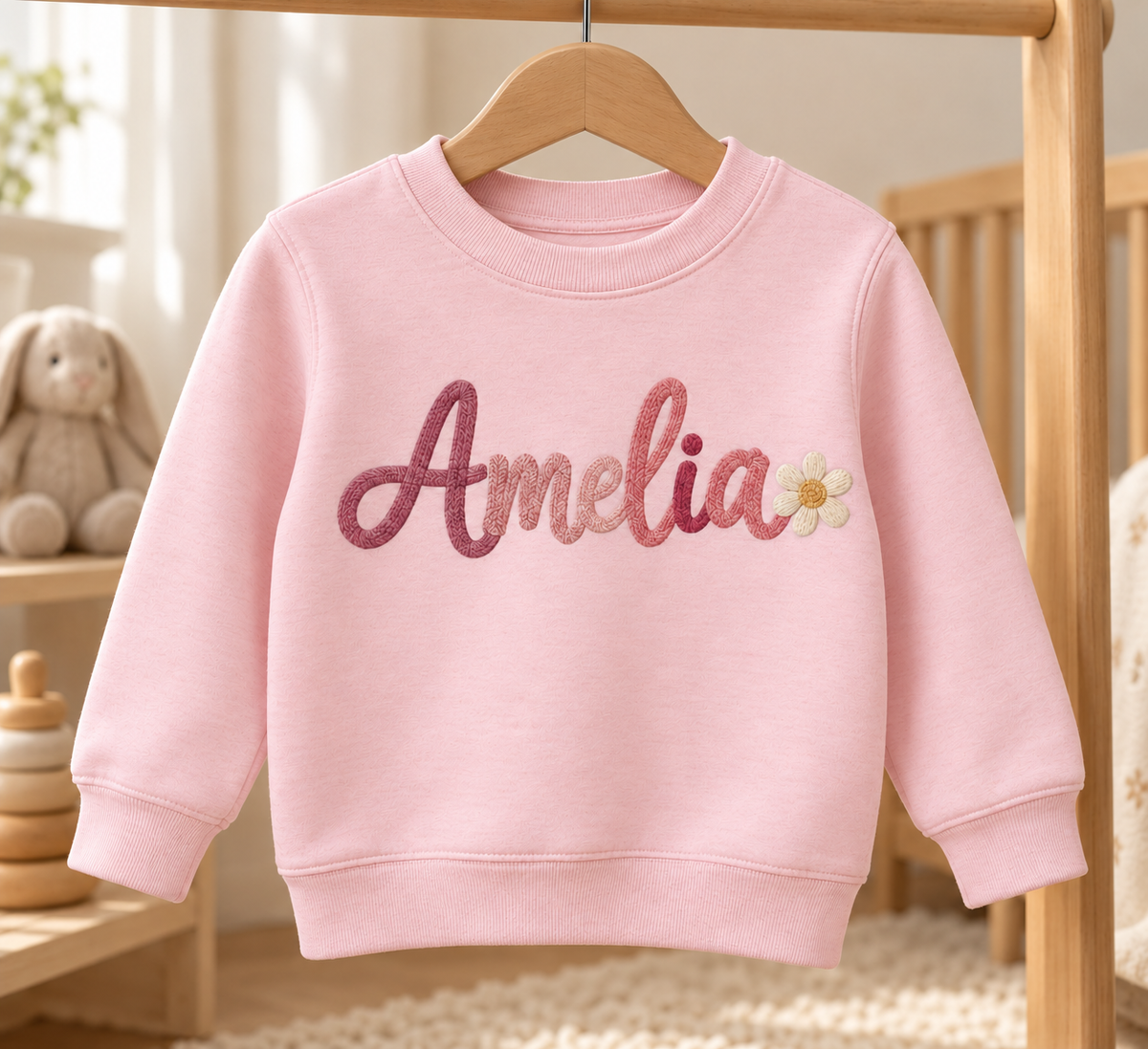 Pink personalized toddler sweatshirt with Amelia sample name and daisy. Flat printed embroidery effect; illustrative mockup.